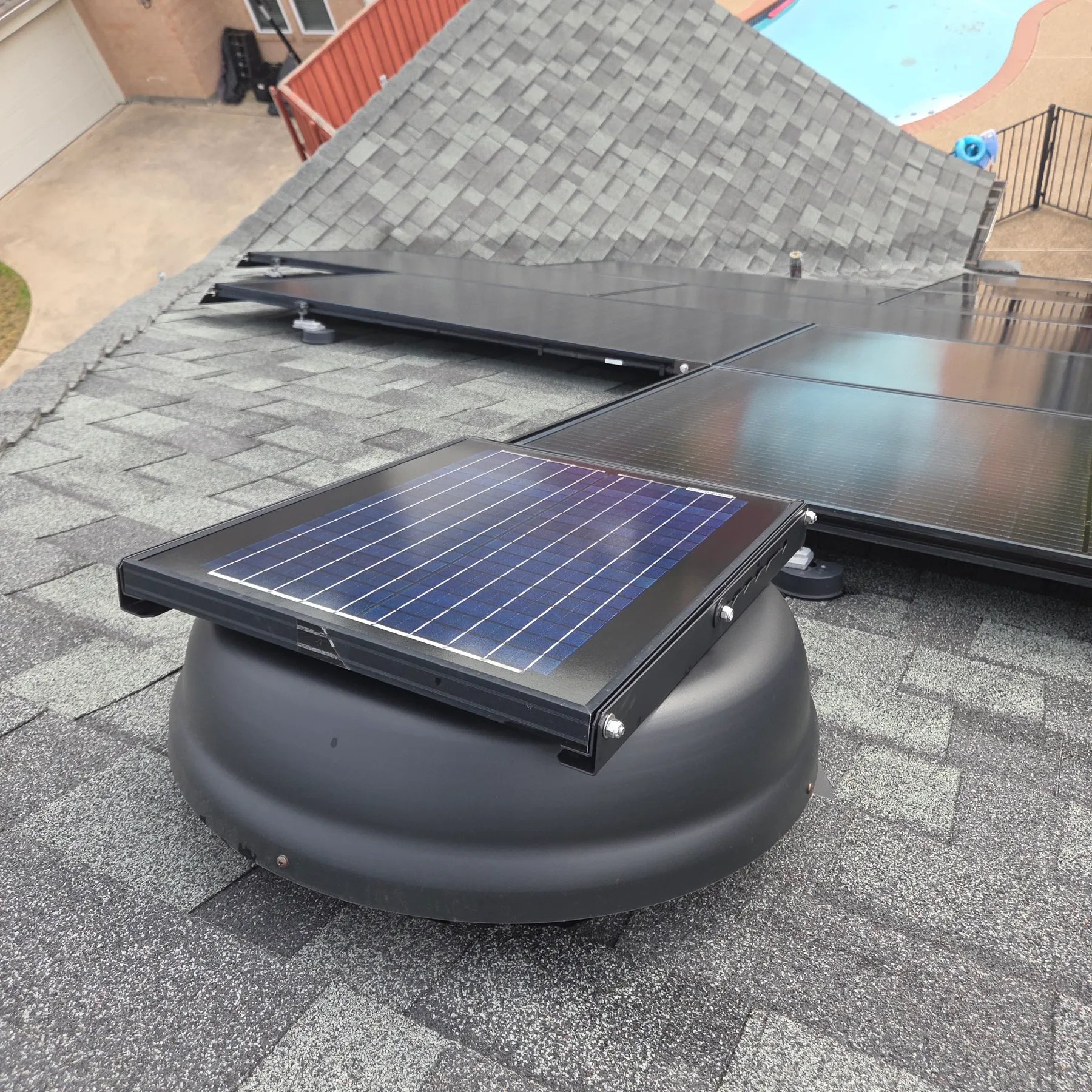 A solar-powered attic ventilation fan mounted on a shingle roof next to a solar array